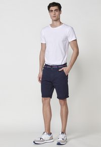 Homme se tenant avec une main dans la poche, portant un t-shirt blanc, un short marine avec ceinture, et des baskets blanches sur fond clair.
