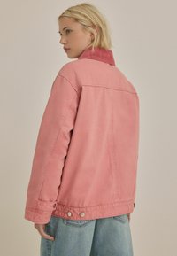 Lyserød denimjakke med en kontrasterende rød corduroykrave, oversized pasform og snap-detaljer foran. Båret over lyseblå jeans.