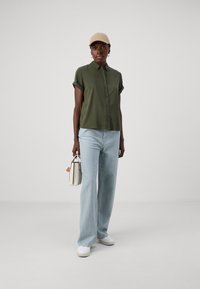 Olivengrønn kortermet skjorte med knappestolpe, lys blå vide jeans, hvite sneakers og en beige caps, holder en liten hvit håndveske.