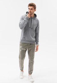 Felpa grigia con tasca a marsupio e cordini, abbinata a joggers olive verdi a gamba stretta con testo bianco. Sneakers bianche completano l'outfit.