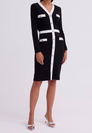 Vrouw draagt een zwarte knielange jurk met witte bies, zilveren knopen, zakken aan de voorkant, lange mouwen en zilveren open teen hoge hakken.