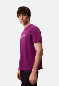 T-shirt en coton violet à manches courtes, col rond, avec logo blanc sur le devant. Texture lisse et coupe régulière.