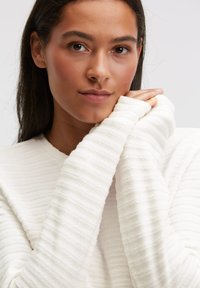 Hellbeiges Langarmshirt mit gerippter Textur. Die Ärmel sind leicht übergroß und verfügen über Daumenlöcher an den Bündchen.