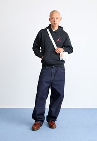 Sort hættetrøje med rødt logo, løstsiddende mørkeblå jeans, brune snørebåndssko og en hvid crossbody-taske med gul accent.