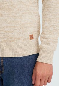Beige gestrickter Pullover mit gerippten Bündchen und Saum, getragen über blauen Jeans, mit einem kleinen braunen Logopatch nahe dem Saum.
