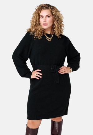 Zwarte gebreide jurk met een riem in de taille, lange mouwen, geribde manchetten en een ontspannen pasvorm. Model draagt gelaagde gouden kettingen en accessoires.