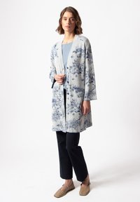 Šviesiai pilkas kimono stiliaus kardiganas su mėlynu kraštovaizdžio raštu, atviro priekinio dizaino, plačiomis rankovėmis ir kelio ilgio kirpimu, derinamas su mėlynu marškinėliais ir tamsiomis kelnėmis.