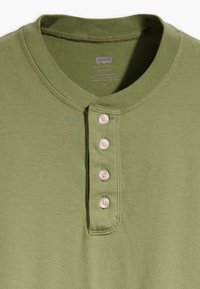 Olivová henley košeľa vyrobená z jemnej látky. Má okrúhly výstrih, päť ružových gombíkov a nenápadné švy pozdĺž okrajov.