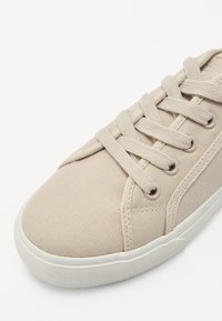 Beige vászon sportcipő, kerek orral, lapos fehér gumitalppal és hat fűzőlyukkal, hozzáillő fűzőkkel. Texturált felület, egyszerű dizájn.