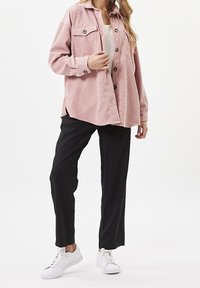 Camicia di velluto a coste rosa con tasche sul petto con bottoni, vestibilità rilassata, indossata sopra una maglietta bianca, abbinata a pantaloni neri e sneaker bianche.