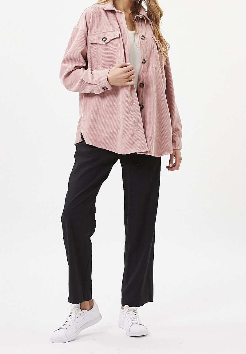 Camicia di velluto a coste rosa con tasche sul petto con bottoni, vestibilità rilassata, indossata sopra una maglietta bianca, abbinata a pantaloni neri e sneaker bianche.