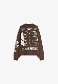 Smeđi oversized dukserica koja sadrži grafičke otiske i tekst. Dugi rukavi, okrugli izrez i rebrasti manžeti. Tekst je istaknuto prikazan na leđima.