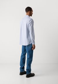 Man die zijwaarts kijkt, gekleed in een lichtblauw geruite lange mouwen shirt, blauwe spijkerbroek en zwarte laarzen, staande op een witte achtergrond.