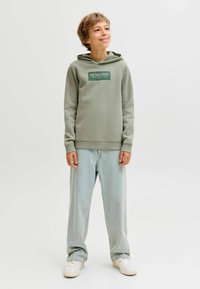 Žalias švarkas su priekiniu logotipu, pagamintas iš medvilnės mišinio, laisvalaikio siluetas, kengūros kišenė, derinamas su šviesiai mėlynomis džinsomis ir baltais sportbačiais.