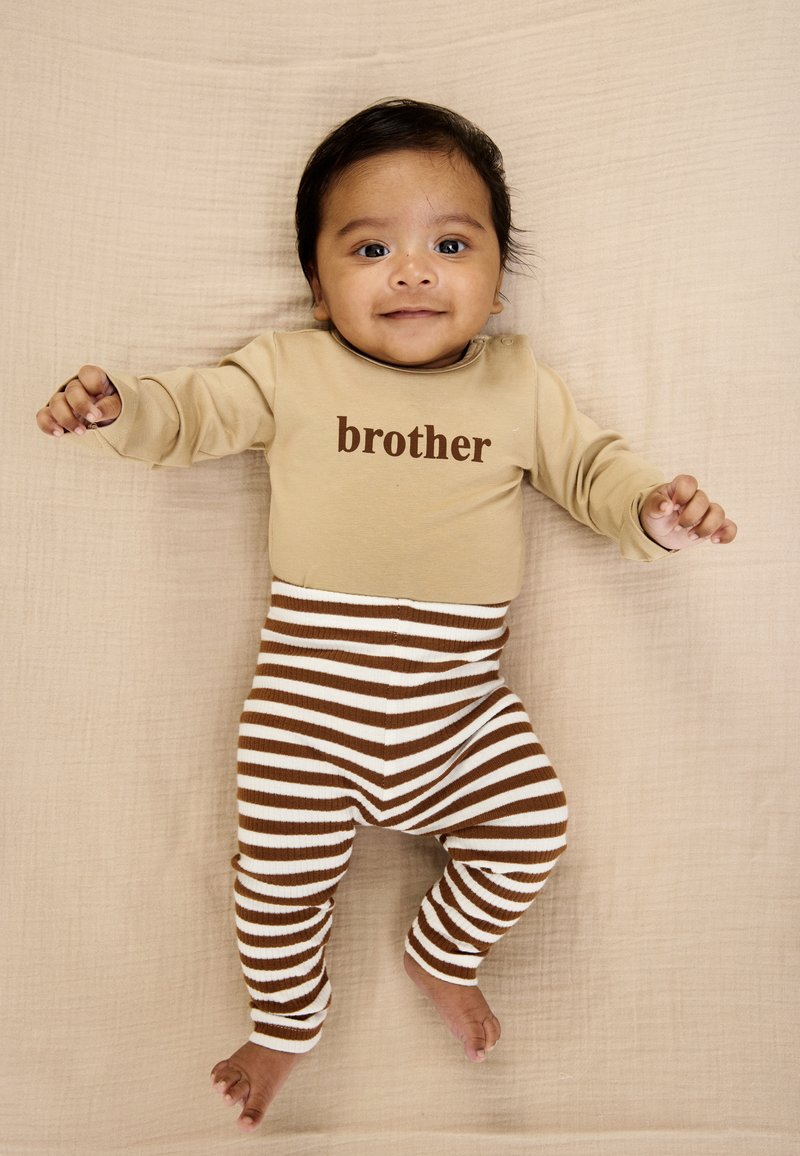 Beige heldækkende onesie med "brother" trykt i brunt, kombineret med brune og hvide stribede leggings. Naturlig tekstureret baggrund.