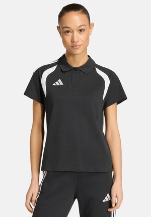 Femme portant un polo noir et un pantalon assorti avec des bandes blanches et le logo Adidas, debout devant un fond clair uni.