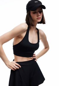 Sort crop top med tykke stropper og en rund halsudskæring, parret med højtaljede sorte shorts. En sort baseballcap med "Basic" logo.