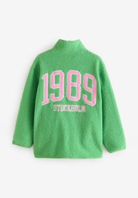 Giacca in pile verde con colletto alto, presenta un grande testo rosa che recita "1989 STOCCOLMA" sulla schiena. Tessuto morbido e vestibilità rilassata.