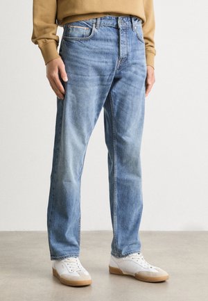 Homme portant un jean droit bleu clair, des baskets blanches et un sweat beige, debout sur un sol gris clair.