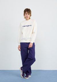 Creme sweatshirt med "carhartt" logo i mørkeblå, parret med løse lilla bukser og lilla sneakers med hvide striber; flad tekstur.