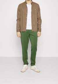 Bruine langemanige shirt, groene broek en witte sneakers. Het shirt is dichtgeknoopt met een borstzak; de broek is goed aansluitend met een gladde textuur.