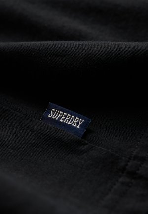 Tela negra con una superficie texturizada que presenta una etiqueta "SUPERDRY" en letras blancas sobre un fondo azul oscuro.