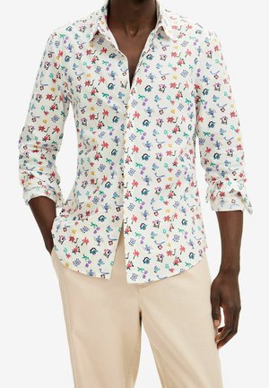 Homme portant une chemise blanche boutonnée avec des imprimés abstraits et floraux multicolores, associée à un pantalon beige clair, mains dans les poches.