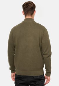 Suéter de punto verde oliva con cuello y puños acanalados, que presenta un diseño texturizado y vista trasera, llevado sobre pantalones negros.