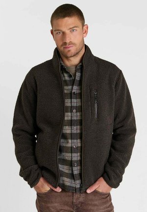 Mann trägt eine dunkle Fleecejacke über einem grau-schwarz karierten Hemd mit den Händen in den braunen Hosentaschen vor einem schlichten Hintergrund.