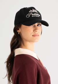 Svart baseballcaps med brodert tekst "Forbudt 10119 Berlin" og bladdesign. Strukturerte form med buet skjerm, båret i en vinkel.