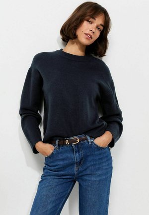 Mujer con un suéter azul marino oscuro y vaqueros azules con un cinturón marrón, de pie con las manos en los bolsillos contra un fondo claro y liso.