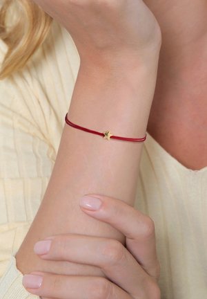 Bracelet en cordon rouge avec un petit pendentif en forme d'étoile dorée. Le cordon est lisse, et l'étoile présente un design à cinq pointes, offrant une apparence délicate.
