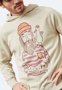 Sudadera beige con un personaje ilustrado que tiene barba, lleva un gorro naranja y sostiene un pájaro. El diseño incluye patrones a rayas y texto.