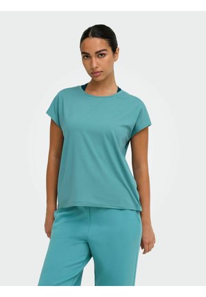 Femme aux cheveux foncés attachés en queue de cheval, portant un t-shirt bleu sarcelle à manches courtes et un pantalon assorti, debout devant un fond clair uni.