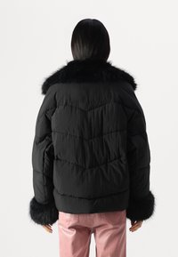 Veste doudoune noire avec col et poignets en fourrure, présentant un design matelassé et une coupe décontractée, associée à un pantalon rose brillant.