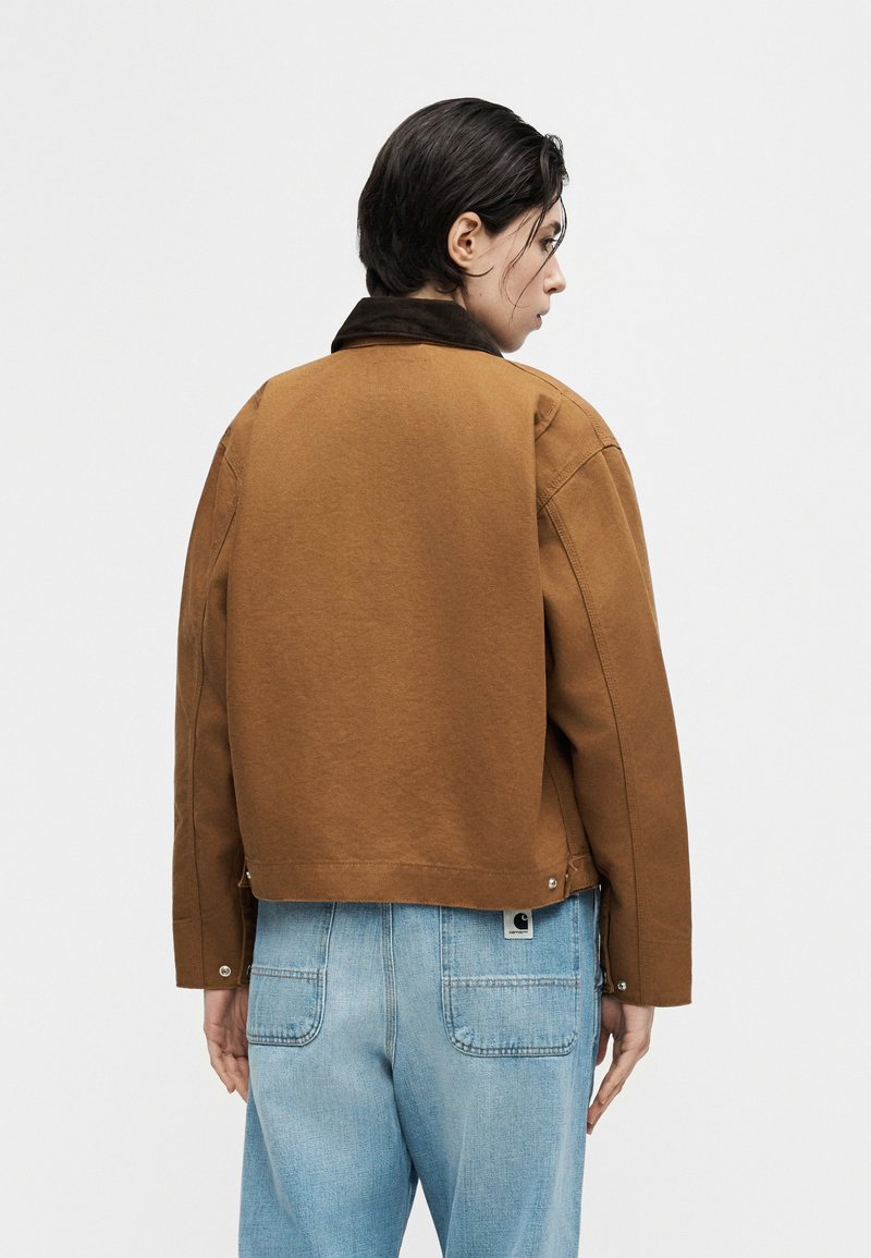 Person with short dark hair wearing a brown jacket with black collar and light blue jeans, shown from the back against a plain background.