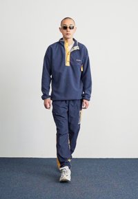 Pull-over en polaire marine avec des accents jaunes et une petite poche. Assorti avec un pantalon marine correspondant, doté d'une bande jaune et d'un patch.