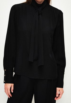 Frau trägt eine schwarze Bluse mit langen Ärmeln, einem gebundenen Halstuch und geknöpften Manschetten, kombiniert mit schwarzen Hosen.