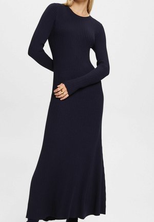 Femme portant une longue robe en maille côtelée bleu marine à manches longues, debout avec une main sur la taille devant un fond uni.
