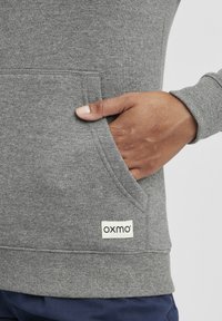 Szürke pulóver elülső zsebbel, texturált anyaggal, bordás mandzsettákkal és hemzsével, valamint egy "oxmo" felirattal a bal alsó oldalon.