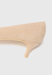 Zapato de tacón alto en ante beige con un tacón bajo y estrecho y una textura suave. Presenta una puntera afilada y un diseño sin costuras en los laterales.