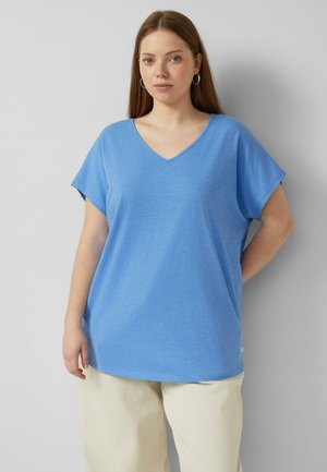 Młoda kobieta z długimi brązowymi włosami, nosząca srebrne kolczyki koła, luźną niebieską bluzkę z krótkim rękawem i dekoltem w serek oraz kremowe szerokie spodnie, stoi przy szarej ścianie.