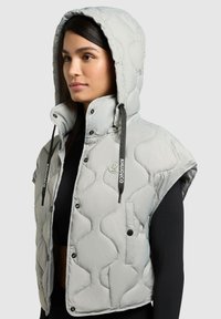 Femme portant un gilet matelassé sans manches gris clair avec capuche, boutons-pression et cordons noirs, par-dessus un haut à manches longues noir, vue de profil gauche.