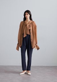 Light brown, textured long-sleeve shirt with a bow tie, paired with dark blue trousers and off-white flat shoes. Simple detailing and tailored fit.
