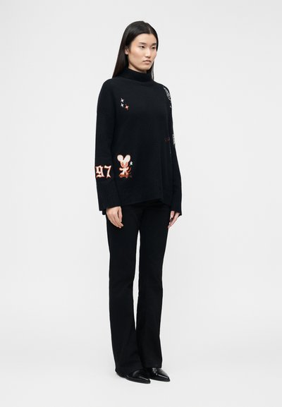 Femme portant un pull à col roulé noir avec des patchs de dessin animé et "97", ainsi qu'un pantalon noir évasé, debout devant un fond clair uni.
