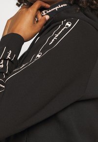 Felpa nera con cappuccio caratterizzata dal logo Champion rosa sul colletto e lungo la manica. Il materiale è morbido, con una finitura leggermente texture.