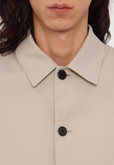 Cămașă bej cu guler, care prezintă o închidere cu nasturi și un interior contrastant de un bleumarin închis. Fabricată dintr-un material neted cu o textură subtilă.