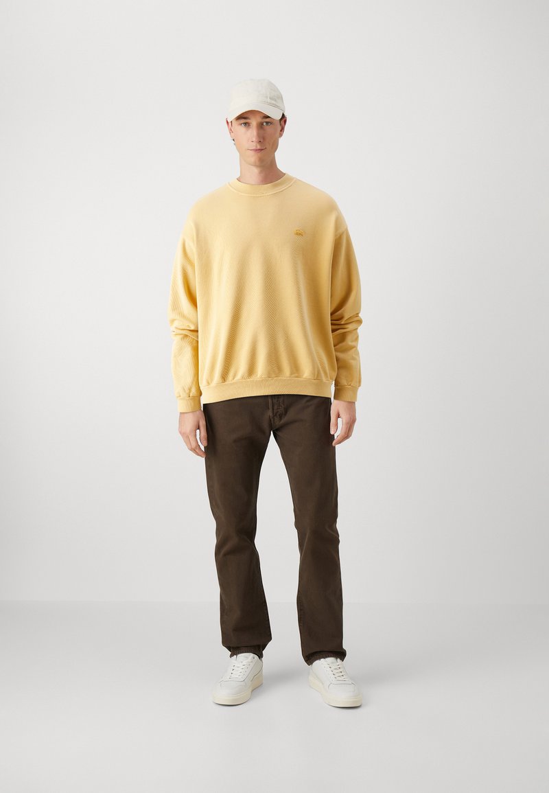 Geel gestructureerd sweatshirt, bruine broek en witte sneakers. Het sweatshirt heeft een ronde halslijn en ribgebreide manchetten. Een lichte pet maakt de outfit af.