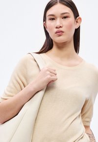 Pulover bej cu mâneci scurte și decolteu rotund. Textură netedă, croială conică. Modelul ține o geantă deschisă la culoare, evidențiind un aspect curat.