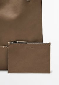 Sacca in pelle marrone caratterizzata da una texture liscia, chiusura con zip e logo impresso. Accompagnata da una borsa in pelle marrone più grande.